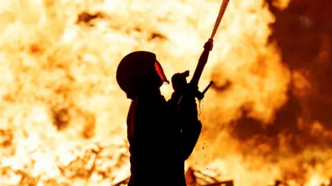 PA Media A firefighter in fireproof gear, including an enclosed helmet, sprays a jet of water from his hose into the air. Behind his flames explode. 