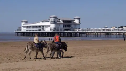 Two people ride donkeys along the beach at Weston-super-Mare. In the background the main structure of the town's grand pier is visible, and the sea is also visible, with the tide about halfway along the pier.