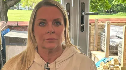 Lisa Barker sits in a living room in front of a patio door. She has shoulder-length blonde hair and is wearing a cream hoodie. Her mobile phone is on the table next to her. 