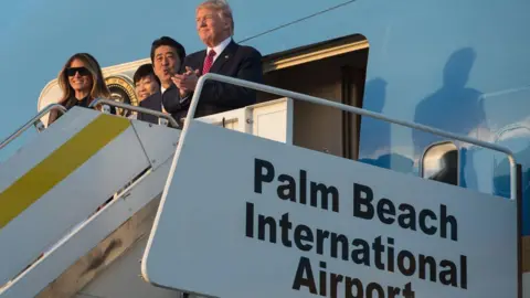 Getty Images Donald Trump and Melania Trump stand atop the stairs of Air Force One. On the side of the stairs, there is a sign that reads Palm Beach International Airport. 