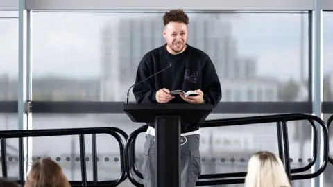 Literature Wales Connor on stage at the Senedd for an event. You can see people are seated that are facing him as guests for the talk