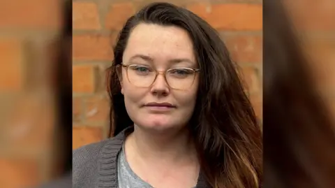 A headshot of a woman with long dark hair and glasses standing in front of a brick wall and looking at the camera.