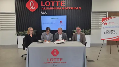 BAL Three men and one woman sit at a long table with company insignia behind them. Two men in the middle have paperwork in front of them and one man is signing papers. The woman and fourth man are smiling at the camera by a signing that says "signing ceremony".
