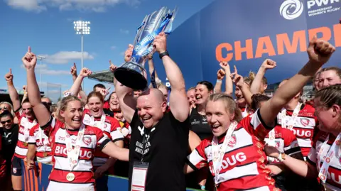 Sean Lynn holds the Premiership Women's Rugby trophy in the air surrounded by the players after winning the 2024 final