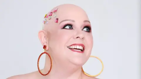 Robyn Law A bald woman smiles as she looks up. She is wearing big gold hoop earrings with red gems on them and has colourful gems on her head. She is wearing pink eyeshadow, false eyelashes and lipstick. 