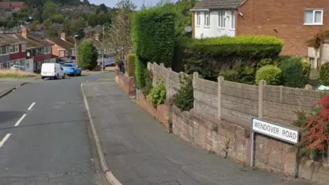 A residential street, with houses either side. There is a white sign with black lettering that reads Wendover Road in the bottom right hand corner of the image. There is a white van and a blue car parked just before a bend in the road to the left hand side of the image.