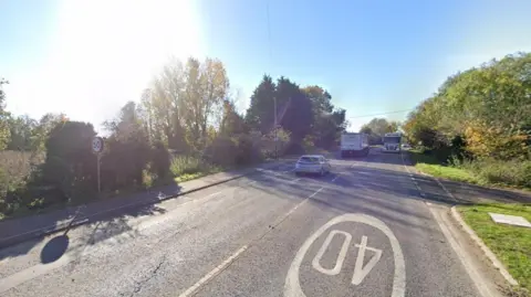 Google view of the A10 road near to Milton. There are lorries and cars on the road and trees at the side. Speed limit signs are at the side of the road and painted on the surface of the road