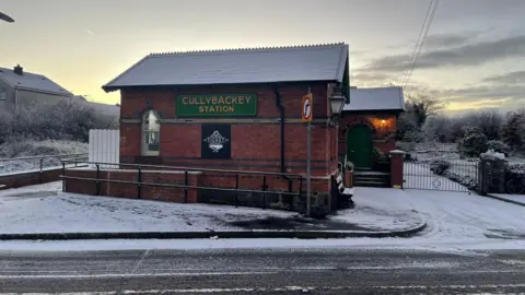 Mark Osmer A red brick building, with a green sign on the front reading "Cullybackey Station". The path in front of the building is covered in snow. 