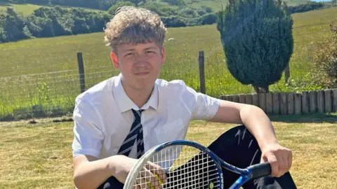 Jack sits in a garden that backs onto a large field in the countryside. He's wearing a school shirt and tie and is holding a tennis racquet.