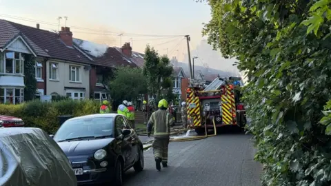 A residential street with a haze of smoke in the sky due a house fire. The street is a narrow lane and has houses on one side and a green bush along the other. Cars are parked in front of the houses and a fire engine is blocking the street. Several firefighters are standing in front of the property and yellow hoses are in the road.