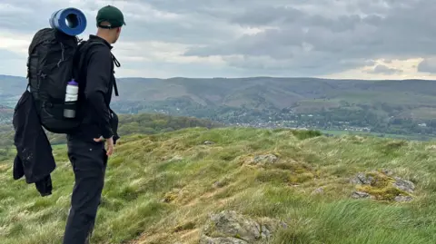 Family A young man with his back to the camera, wearing a green baseball cap and a black top and black trousers. On his back is a rucksack with a bedroll strapped to the top. He is looking out over a green valley.