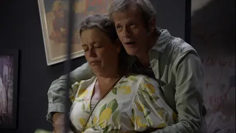 Two actors, a man and a woman on stage. The man is holding the woman in his arms and she looks upset.