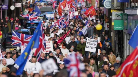 Crowds walk down the road in a protest waving Union flags. A banner says "detain, process, deport". There are shop signs to one side of the town centre road.