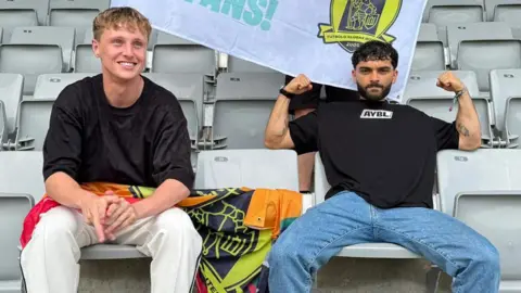 Ellis Platten/Alfie Indra Ellis Platten and Alfie Indra sitting next to each other on light grey seats inside a stadium. Platten has a black T-shirt on and his smiling. Indra also has a black T-shirt on and is flexing his arms.