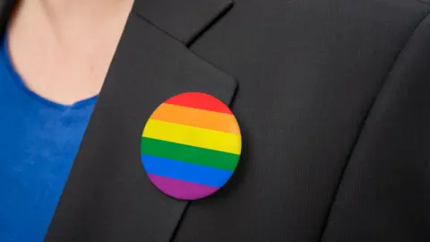 A round badge of rainbow colours in horizontal stripes, pinned to the lapel of a black suit jacket 