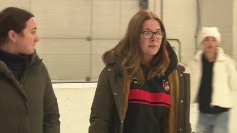 Hannah Driver (right) skating on the ice she's wearing a green coat over a black hoody. She has long hair and glasses. Another skater wearing a white jacket and hat can be seen in the background.