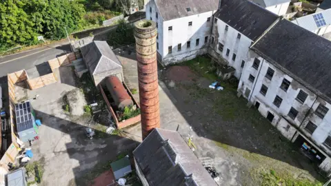 SOSE An aerial view of the old mill site with white main buildings and a large brick chimney stack
