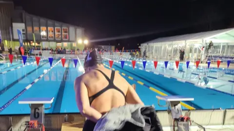 Frozen Ferns A swimmer disrobing ready to get into the pool. It is dark and there is some floodlighting.