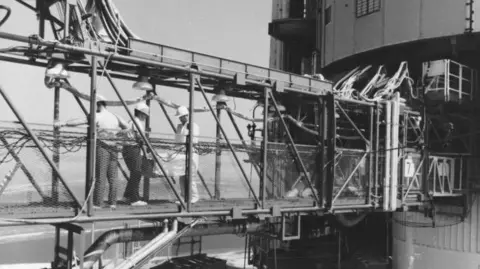 NASA/ESA Keith Wright carries the Lunar Laser Ranging experiment across the swing arm towards the Apollo 11 Saturn V with two other men in hard hats.