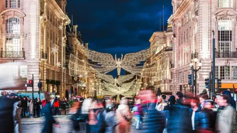 Crowds cross a busy West End street at night beneath large angel-shaped Christmas lights suspended between illuminated buildings.