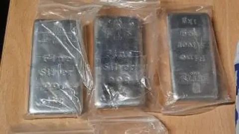 Three silver bars in clear plastic bags, lined up on a wooden desk.