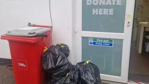 A pile of black binbags leaning against a red plastic wheelie bin, outside a white UPVC door with a "Donate Here" and a fire exit sign.