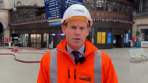 Network Rail Network Rail Scotland's route director Ross Moran, wearing a white hard hat and and orange hi-vis jacket stands on the concourse of Glasgow Central station with fire hoses lying on the floor and tape cordons behind him. 