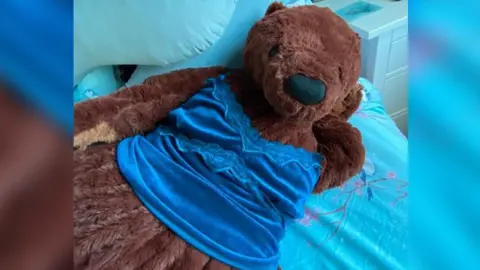 PA Media The teddy is brown, and has a black nose and eyes. It is laying on a blue bed, and is wearing a dark blue top. 