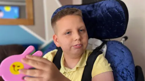 A boy with short brown hair, wearing a yellow polo shirt, is sat in a blue wheelchair. He is holding a plastic toy in the shape of a hand