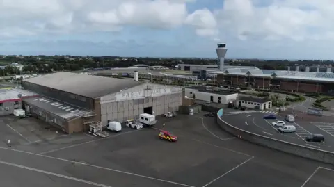 Large grey hangar building with old sign where some of the letters are no longer visible. Air control tower and other airport buildings visible in the background