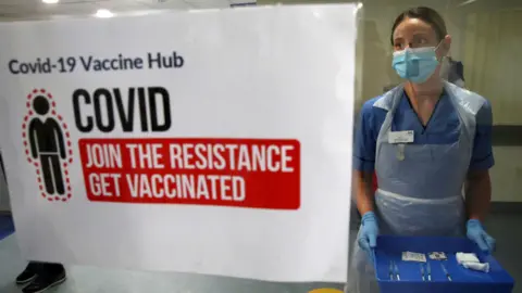 PA Media A woman in a blue uniform and mask carrying a tray for a vaccination session. She is next to a sign about getting vaccinated against Covid