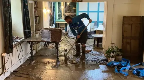 A man in a blue top, wearing boots and holding a broom clears debris from the floor of a flooded pub after the water has receded.
