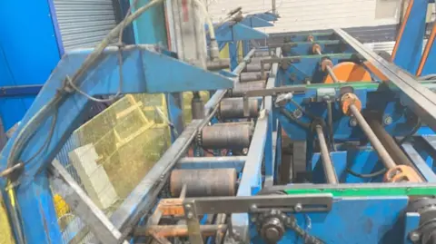 HSE A blue machine with open cogs and chains. There are various metal components which are unscreened. The machine has a row of rollers and other workings. There are unpainted metal shutters in the background.