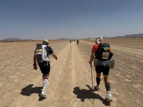 The backs of two men walking through the Sahara desert surrounded by dry land, carrying rucksacks on their backs, one walking with a stick