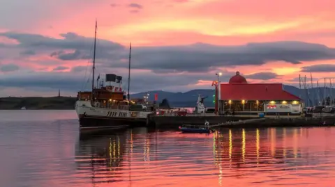 The Waverley paddle steamer - a boat with red and black chimneys and two masts - docked in a harbour. There are boat masts and a red-roofed building at the harbour. The sky is pink and orange. 