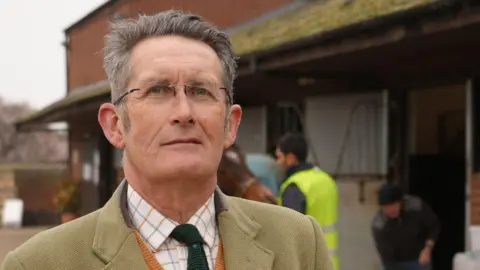 Kevin Needham is standing on the left in front of a horse stables. He has short grey hair and is wearing glasses, a white chequered shirt, green tie and a tweed coat.
