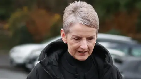 A close-up image of Lady Eleanor Donaldson as she walks out of court. She is an older woman with short, grey hair and is looking down. She is wearing a black, puffer raincoat and a pair of silver stud earrings.