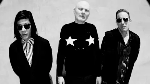 A black-and-white photo of the band The Smashing Pumpkins. They are all wearing dark outfits and two of them are wearing sunglasses.