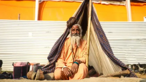 Ankit Srinivas Baba Amarnathji, a 60-year-old monk wearing saffron robes, sits outside a small tent he has set up with cloth and plastic sheeting atop three bamboo poles.