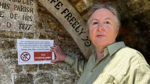 Julia Gregory / BBC A woman stood next to a stone wall with inscriptions on it. She is holding a sign reading "Not drinking water".