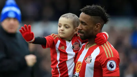 Former England Striker Jermain Defoe, holding Bradley Lowery. The pair are wearing red and white striped Sunderland AFC strips, Bradley is also wearing some bright red SAFC gloves.