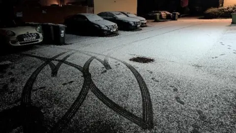 Lucie Johnson Two love hearts have been made in a light dusting of snow on a road with cars parked behind. It's a dark scene taken early in the morning. Footsteps can also be seen in the snow.