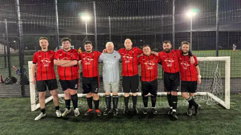 Man v Fat A row of eight men lined up, posing for a group picture in front of a white football goal at night. There are bright overhead lights shining down onto the pitch. The men are all wearing black shorts and red shirts, except for the goalie in the centre wearing an all grey uniform.