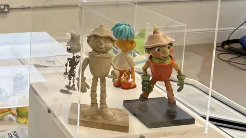 Plasticene figure of a flowerpot man with hat and terracotta pot top next to a painted version in a display case at the exhibit.