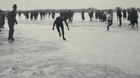 Michelle Bullivant A black and white image of two skaters racing on a frozen field. Spectators can be seen behind them. On their right is a police officer, who is also on skates.