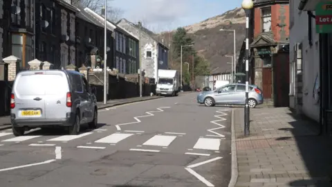 A quiet street scene in Ynyshir. Terraced buildings line both sides of the road, including older brick structures and houses with dark stone façades. Several vehicles are visible: a silver van travelling along the road, a small light‑coloured car parked on the right side near a junction, and a larger white van further ahead.