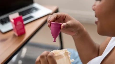 Focus Pixel Art/ Getty Images A woman to the right holding a pink menstrual cup in her hand. She is holding a small fabric pouch in her other hand. In the background a opened box can be seen on a table, stood next to an open laptop.
