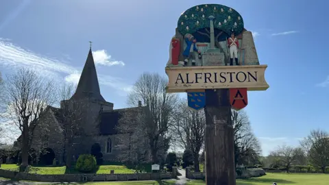 George Carden/BBC A sign which says Alfriston on a wooden post. St Andrews Church is in the background in front of a village green and has a large central steeple in the centre
