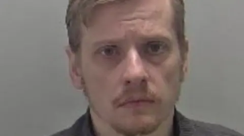 Mugshot of a man with short blonde hair, in a side part with a short fringe. He has a moustache and beard and is wearing a grey shirt.