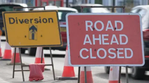 Two road signs. The first is red and reads 'Road closed ahead' while the one behind it is yellow with a black arrow pointing up and says 'diversion'. There are orange and white cones down the central reservation of the road and a queue of cars on the right of them.